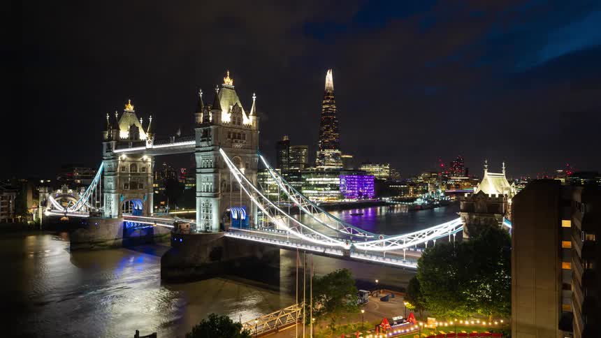 A time lapse of a bridge lit up at night with boats traveling under it with a city lit up in the background