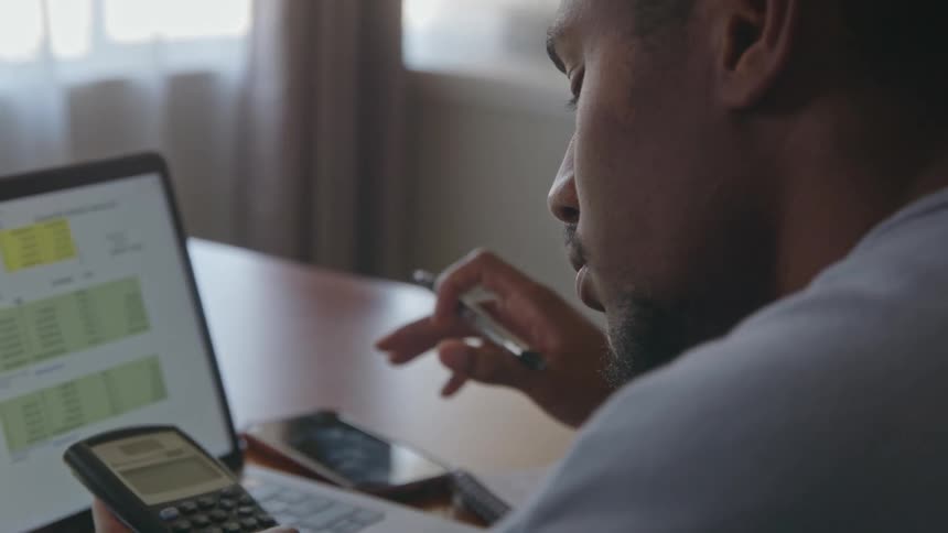 A clsoe up of a mans face as he holds a calculator while looking at a computer screen before writing with a pen