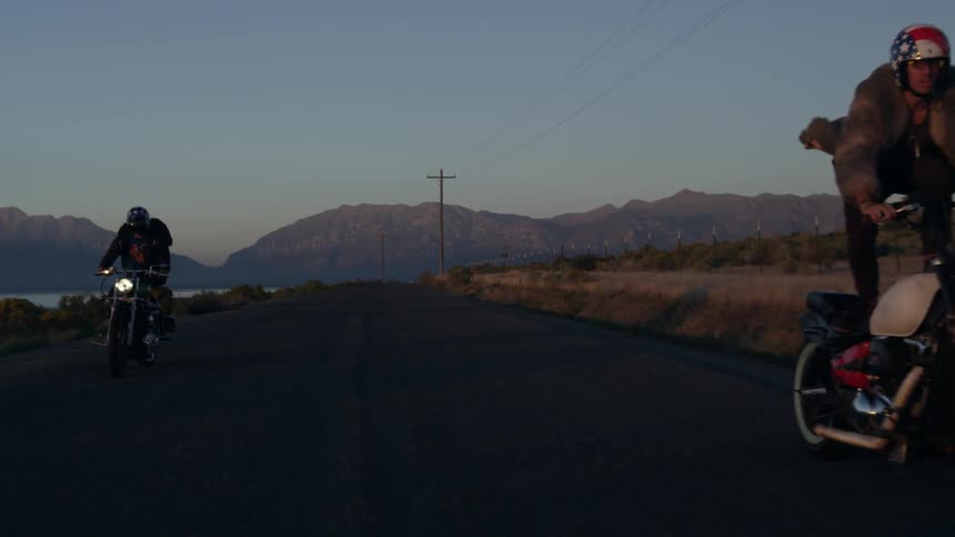 Two motorcycle riders stand on their bikes as they drive down a road