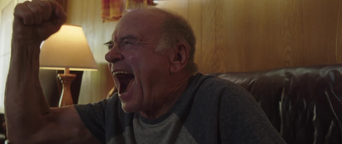 An elderly man sitting on a couch gets excited while watching tv
