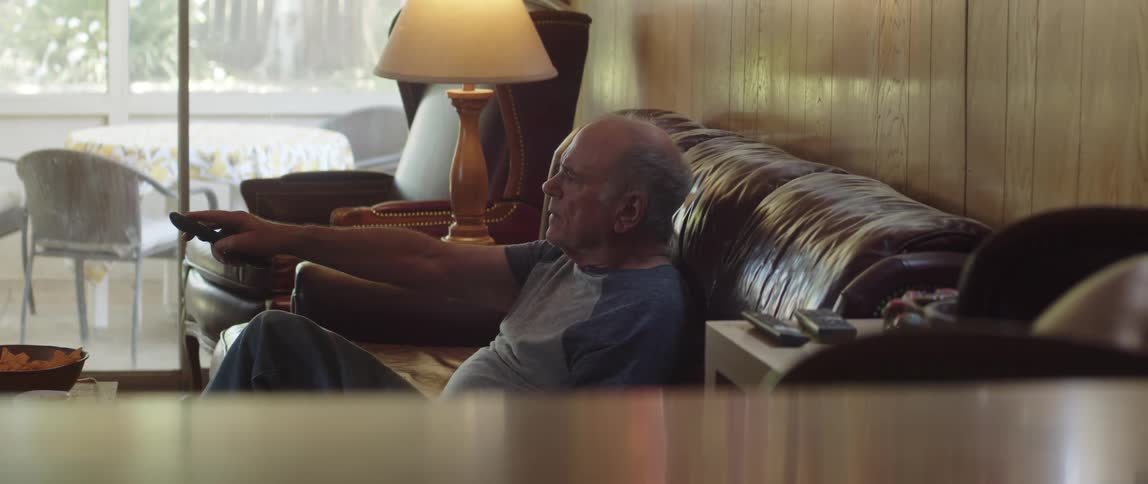 A man uses a remote control to change the channel of a telelvision while sitting on a sofa in the living room of a house during the day