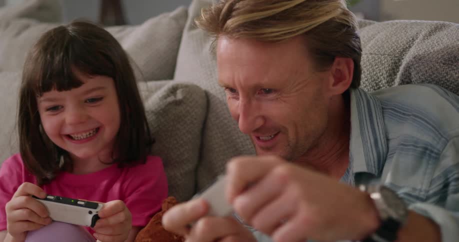 A man plays a video game with a little girl while sitting beside her on a sofa