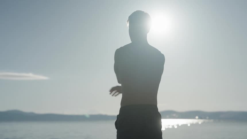 Shirtless man standing in front of the sea stretching his arms while admiring the landscape on a sunny day