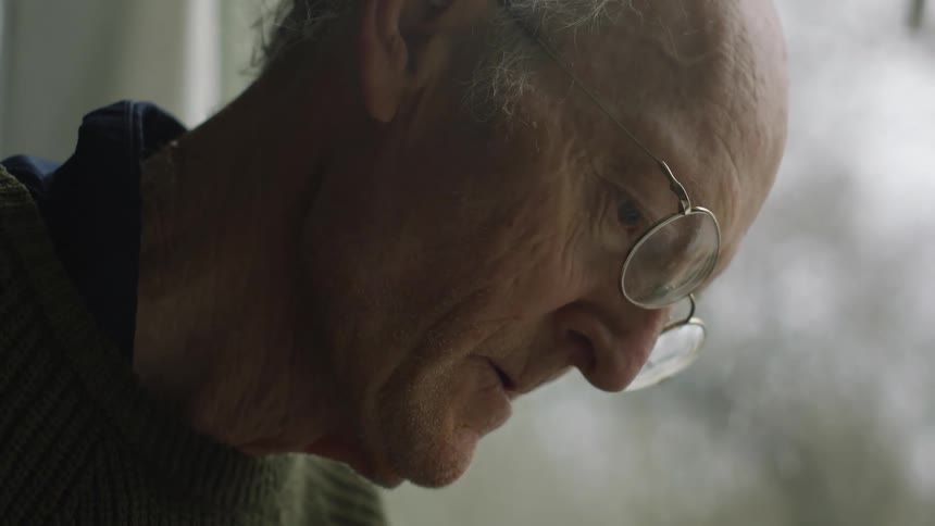 Elderly man wearing a sweater and glasses attentively looking down at something