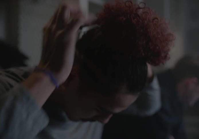 A man with curly hair seated in the dressing room putting his hair up in a bun