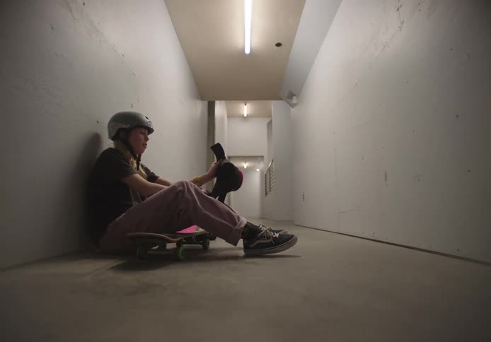 A female skateboarder sitting against a wall in a corridor puts on her helmet and knee pads