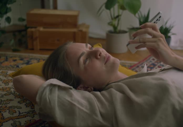 A woman talks into her smartphone while resting on a pillow on the floor