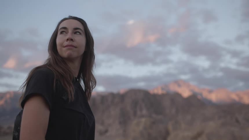 A woman stands outdoors and looks around at the mountainous landscape nearby