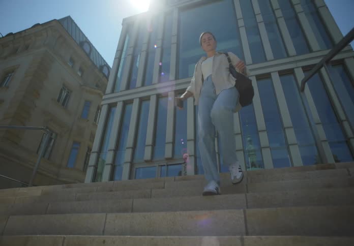 A woman in a hurry with a bag and smartphone in hand walking down the staircase of a large building under a sunny sky