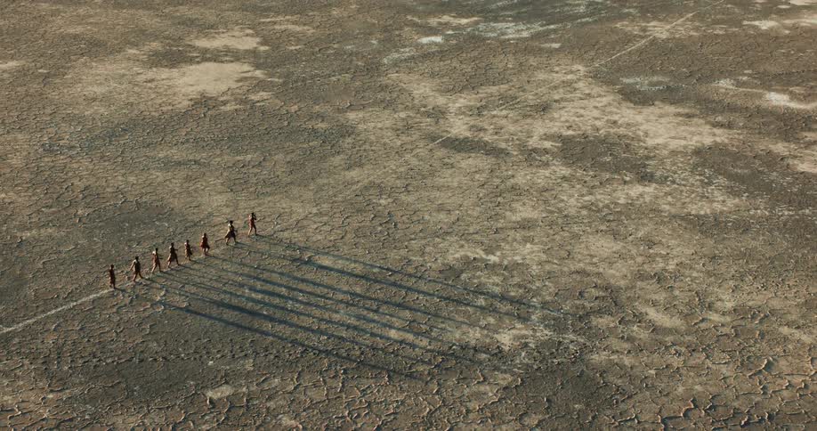 Aerial footage shows a group of bushmen walk in a line through a barren plain