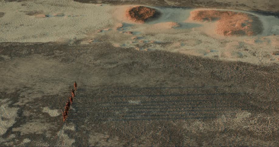 Small group of bushmen walk single file across arid sand casting long shadows