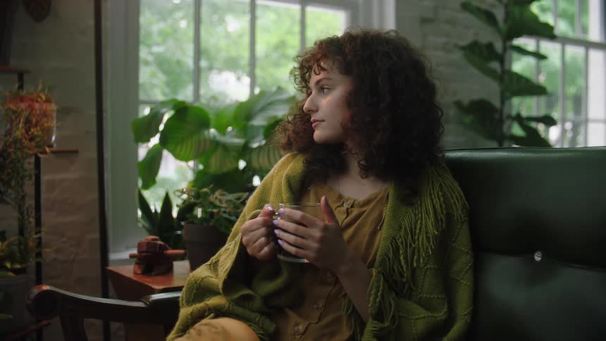 A young woman with curly hair sits on a couch while holding a glass mug
