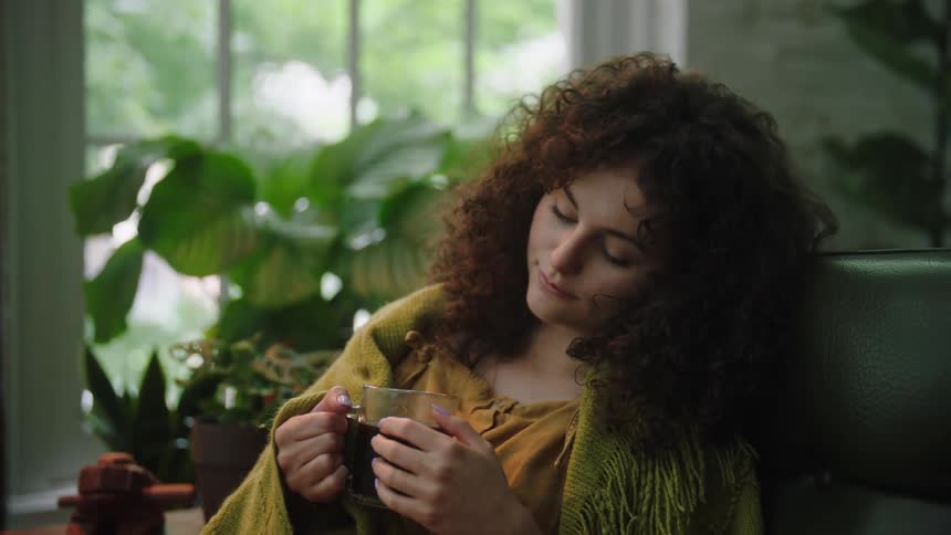 Woman with curly hair sits peacefully cradling her tea in her hands lost in thought a faint smile graces her lips reflecting a moment of quiet contentment and reflection