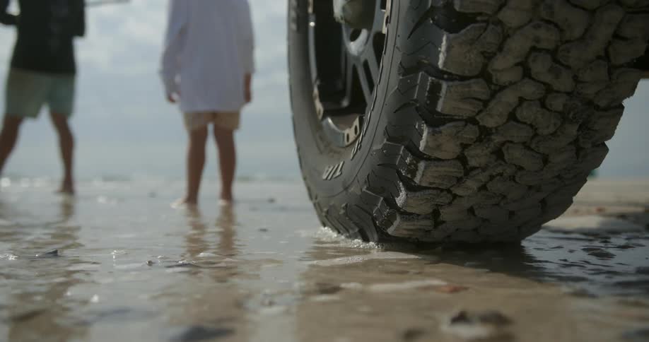 Three people walk on the beach that a nearby vehicle is parked on