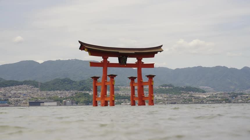 On a cloudy day waves move past torii gate sitting in water a distance from town on shore