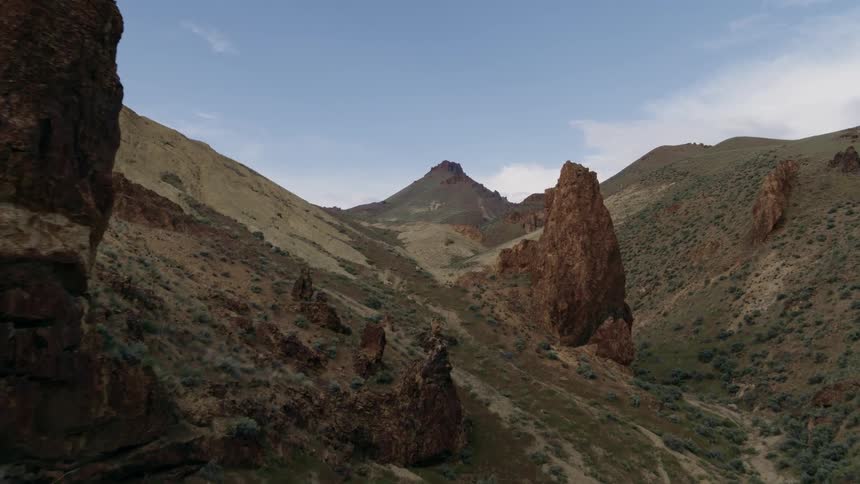 Flying past rock spires in an undulating green valley with a conical mountain peak in the distance