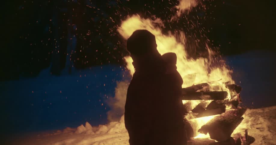 Man with hat and coat is silhouetted against bonfire