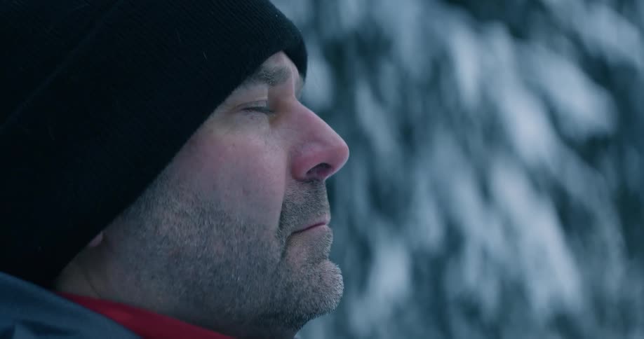 A white man wearing winter clothing is looking up while standing in the cold thinking as he brings his head down is disappointment
