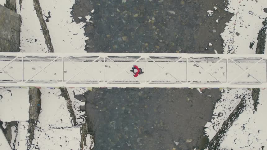 A person wearing winter gear walks over a river via a snow covered bridge