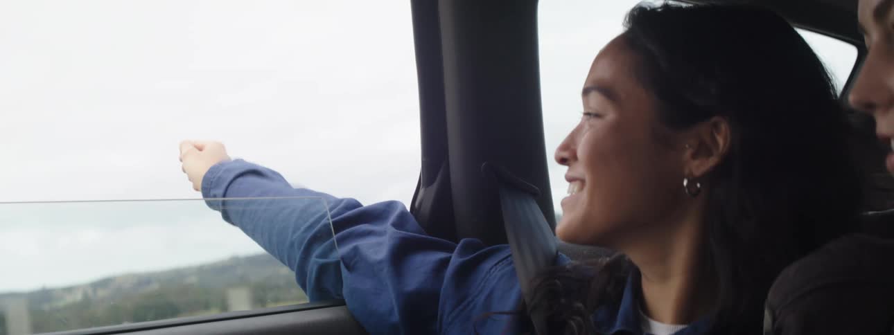 Two young women drive past a grassland and passenger hangs her arm out the window and talks and points