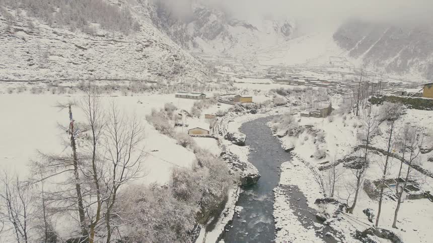An aerial view of a snow covered landscape with a river in a mountain valley and a few buildings