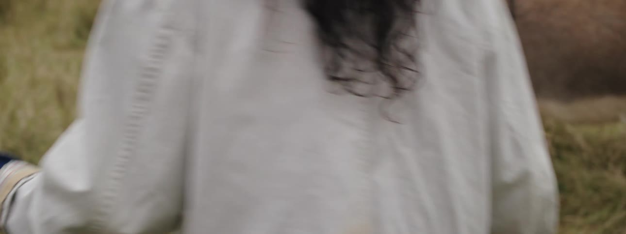 Woman with white jacket and wavy hair approaches donkey to touch it in the meadow