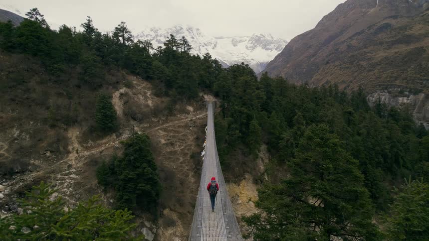 A lone hiker walks across a suspension bridge in a forested area