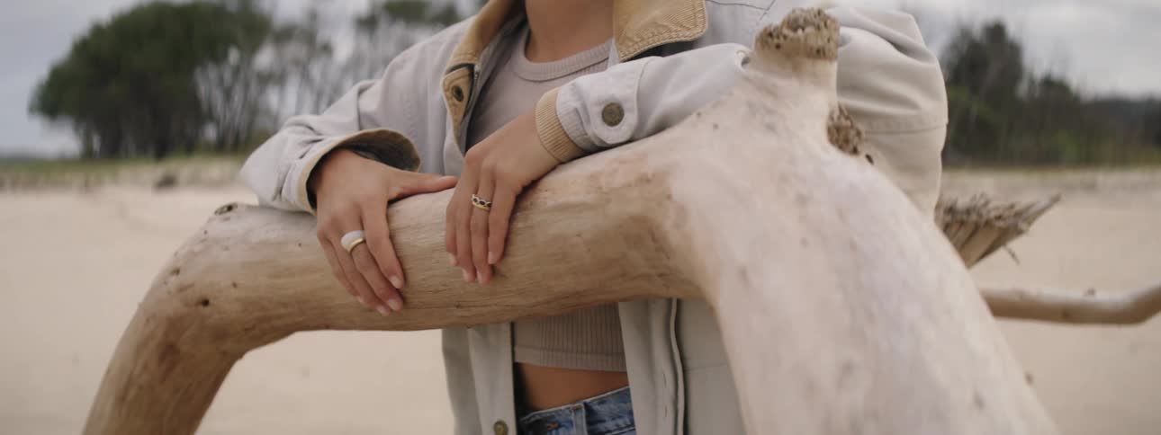 Woman with earrings dark hair and jeans leans against the fallen tree on the beach on a cloudy day