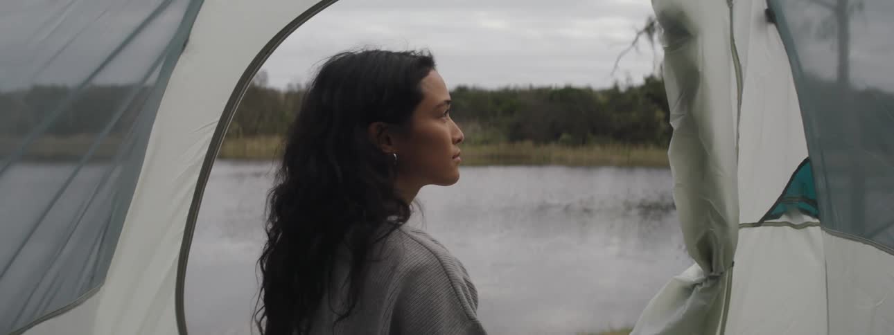 A woman sits in her tent pitched along a lake