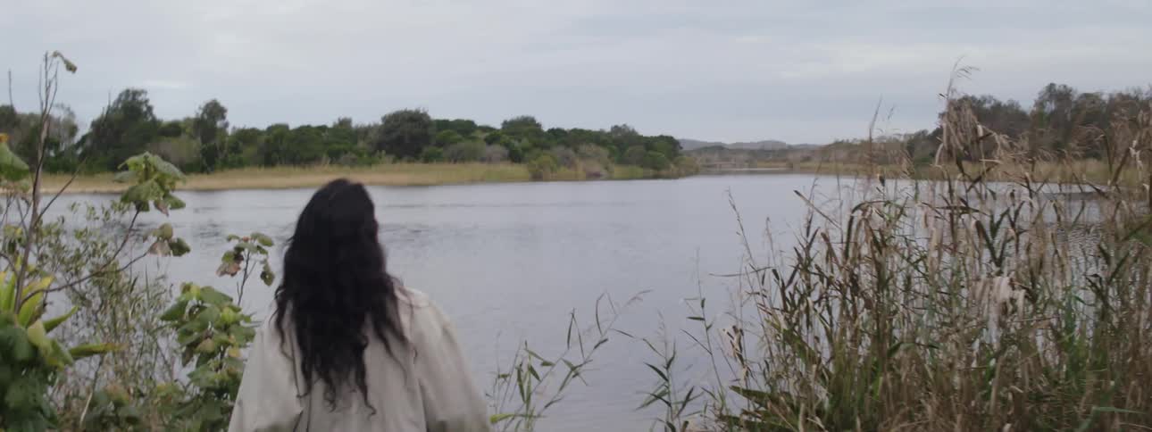 Woman runs through woods onto lakeshore and stops and looks around