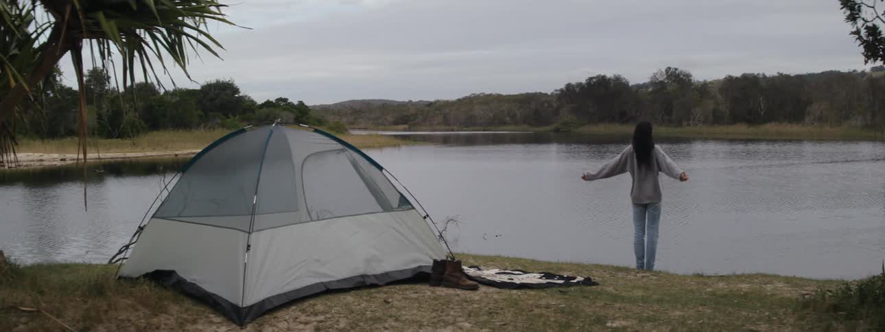 A woman is standing next to a camping tent set up in an area with grasses close to a river