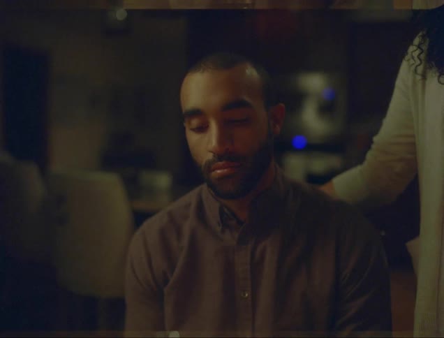 Man contemplates birthday cake with lit candles then smiles toward woman standing near his chair