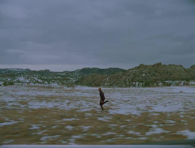 A person running in a grassy field with snow with a mountain range the sky and clouds in the background