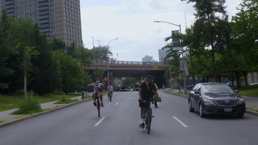 Group of people riding bikes in the middle of the road as they perform stunts with a black sedan car driving along