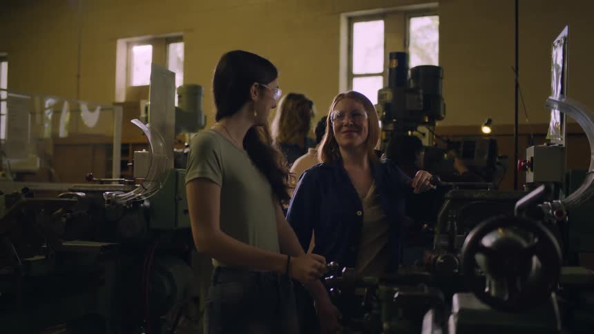 A young woman in a workshop turns a crank as another employee turns a button on a computer to show her how something is done