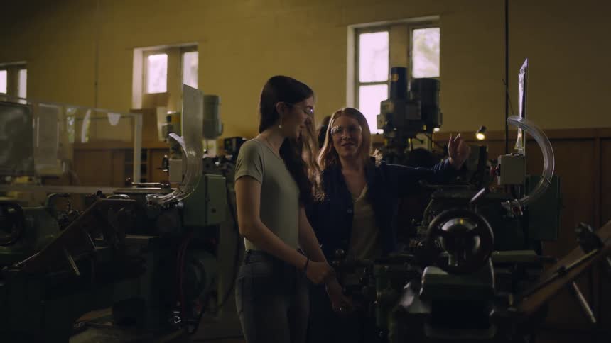Excited older blonde woman wearing protective goggles showing a machine to a younger woman wearing glasses indoors during day time