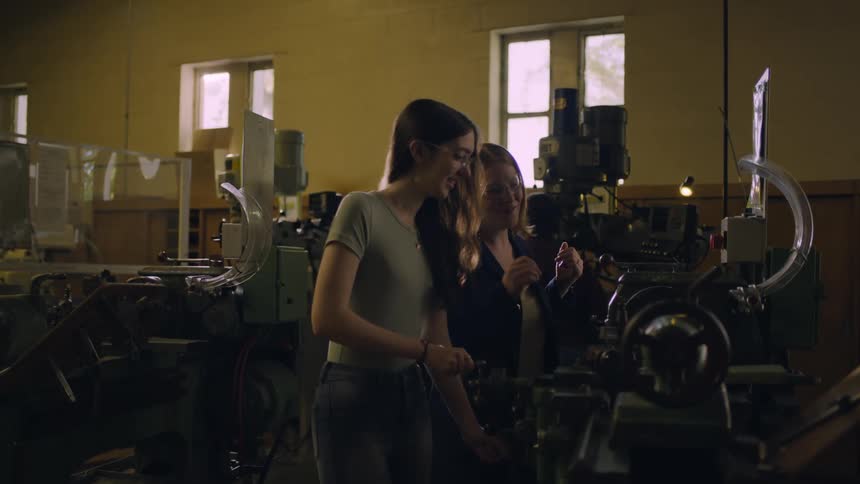 One woman guides another through using machine in workshop