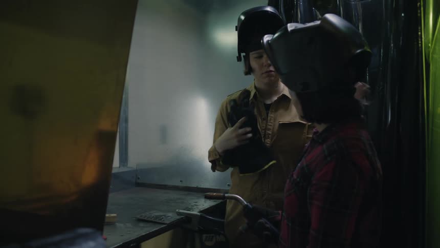 Two female welders stand together while one observes the other