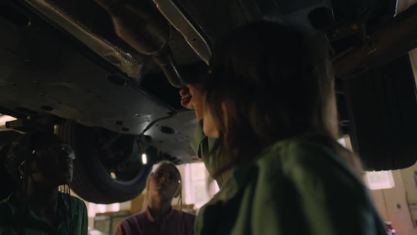 Teacher explains car mechanics to students under a car lift in a workshop