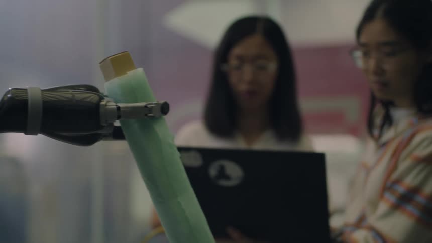 Three women operating a robot from a computer as the robot raises a stick which one of the women takes while wearing a smile of satisfaction