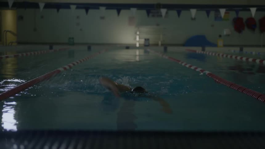 Teenage female swimming straight down a lane in an indoor pool taking her googles up and looking up when she reaches the wall