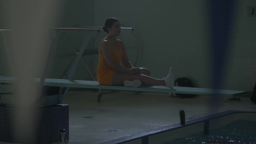 White woman on a yellow bathing suit sitting on a diving board whistling for other people on a pool to start swimming