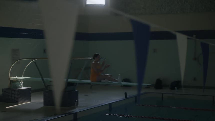 A female sitting on a diving board while two swimmers are swimming in a pool