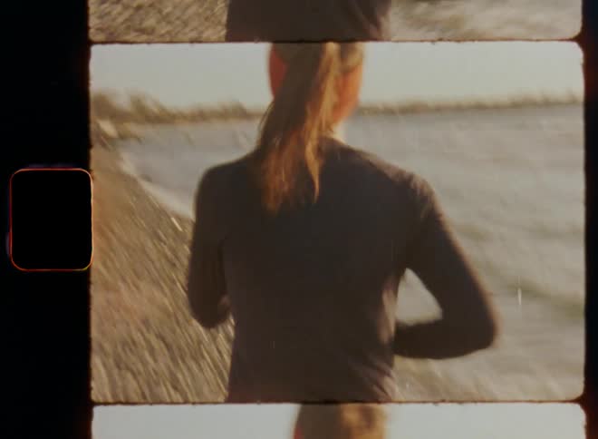 Shaky and dynamic handheld 8mm film footage following a woman runner with brown hair ponytail red headband and black shirt that jogs on the gravel beach training for a marathon competition in a sea bay during daytime