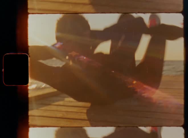 Handheld dynamic 8mm film footage of woman athlete wearing running shoes and doing bicycles while working out on landing bridge in harbour at large lake in the morning sun as the sun rises causing bright lens flares between her moving legs and knees