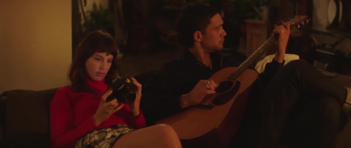 A man plays the guitar while sitting on the sofa next to a woman examining a camera
