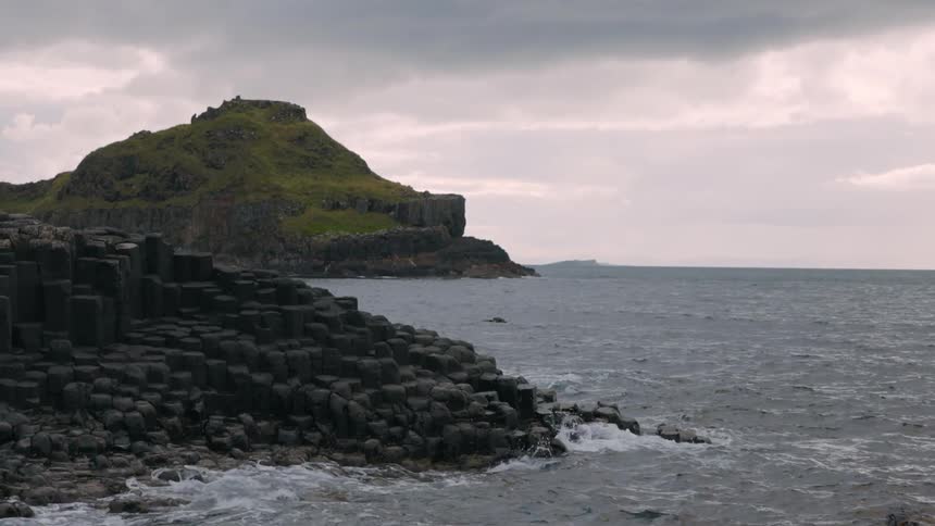 Waves crashing gently onto the shore both on block shaped rocks and a grass covered hill