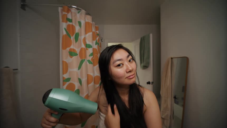 A young woman smiles as she adjusts her hair and dries it with a hairdryer