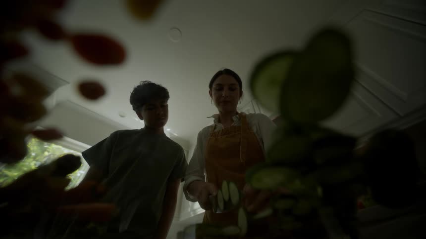 Mother with apron and white shirt cuts vegetable next to son watching in the kitchen