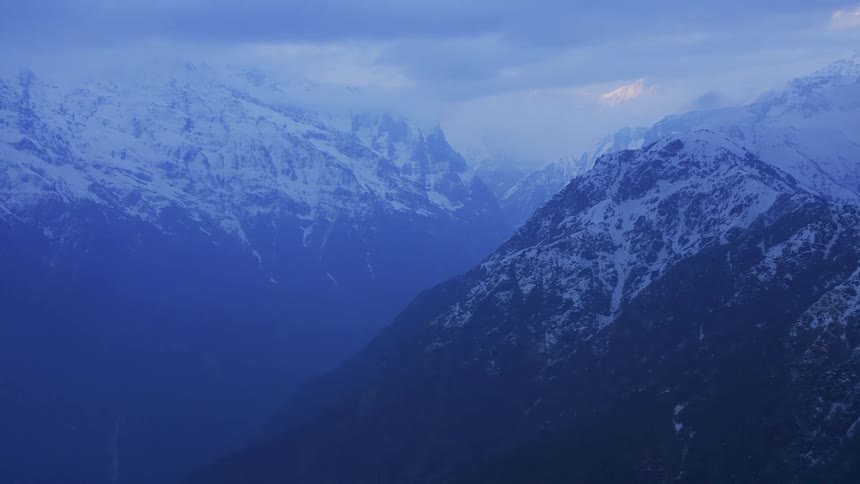 A large snowy mountain range with blue coloring under a cloudy sky